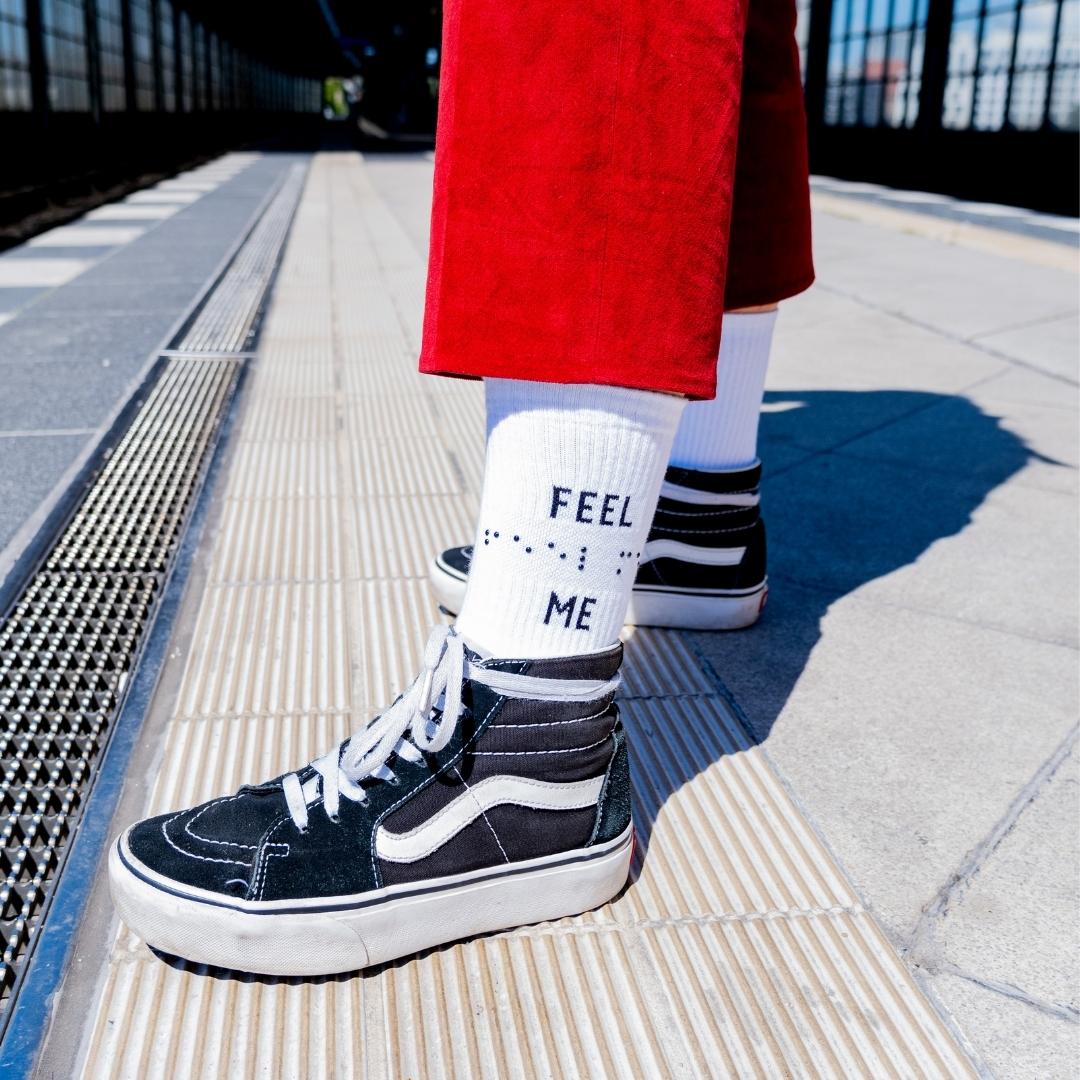 Man sieht zwei Beine mit einer roten Hose, weißen Tennissocken und schwarzen Sneakern. Im Hintergrund sieht man einen Bahnhof. Auf der weißen Socke steht das Statement FEEL ME. Zwischen den zwei Wörtern ist die Botschaft in Braille mit Kuppen aufgepresst.