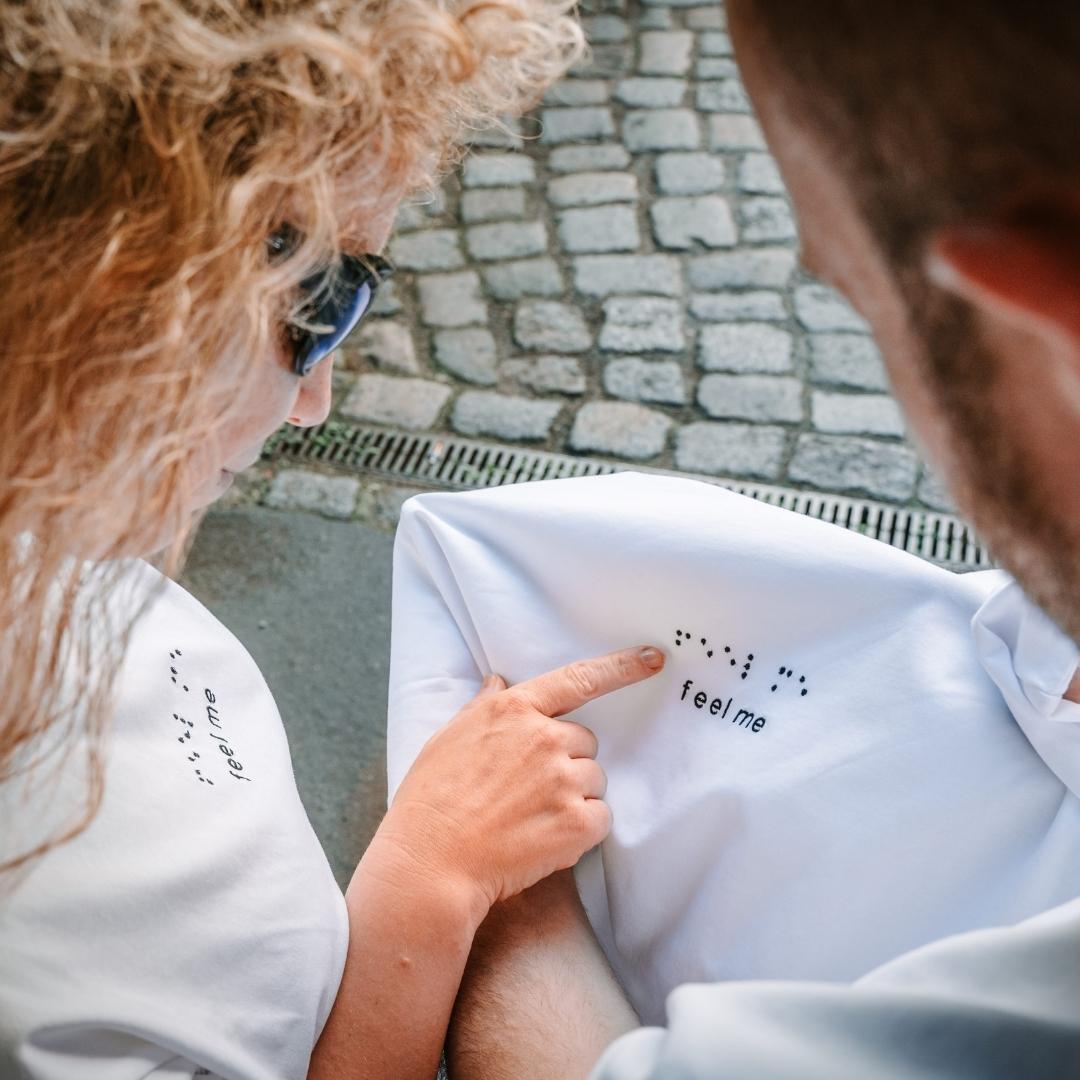 Zwei Personen betrachten das weiße Feel Me T-Shirt, die Frau streicht über das in Brailleschrift geschriebene Statement