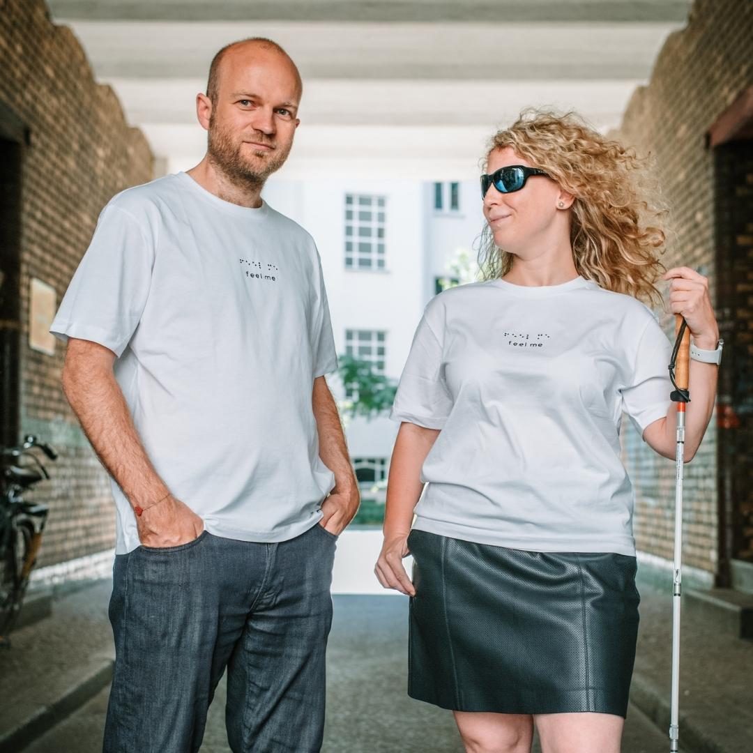 Ein Mann (links) und eine Frau (rechts) tragen beide das weiße Feel Me Shirt. Ihr Blick ist auf ihn gerichtet, sie hält einen Blinden/Langstock in der Hand und trägt eine Sonnenbrille. Er schaut Richtung Kamera.